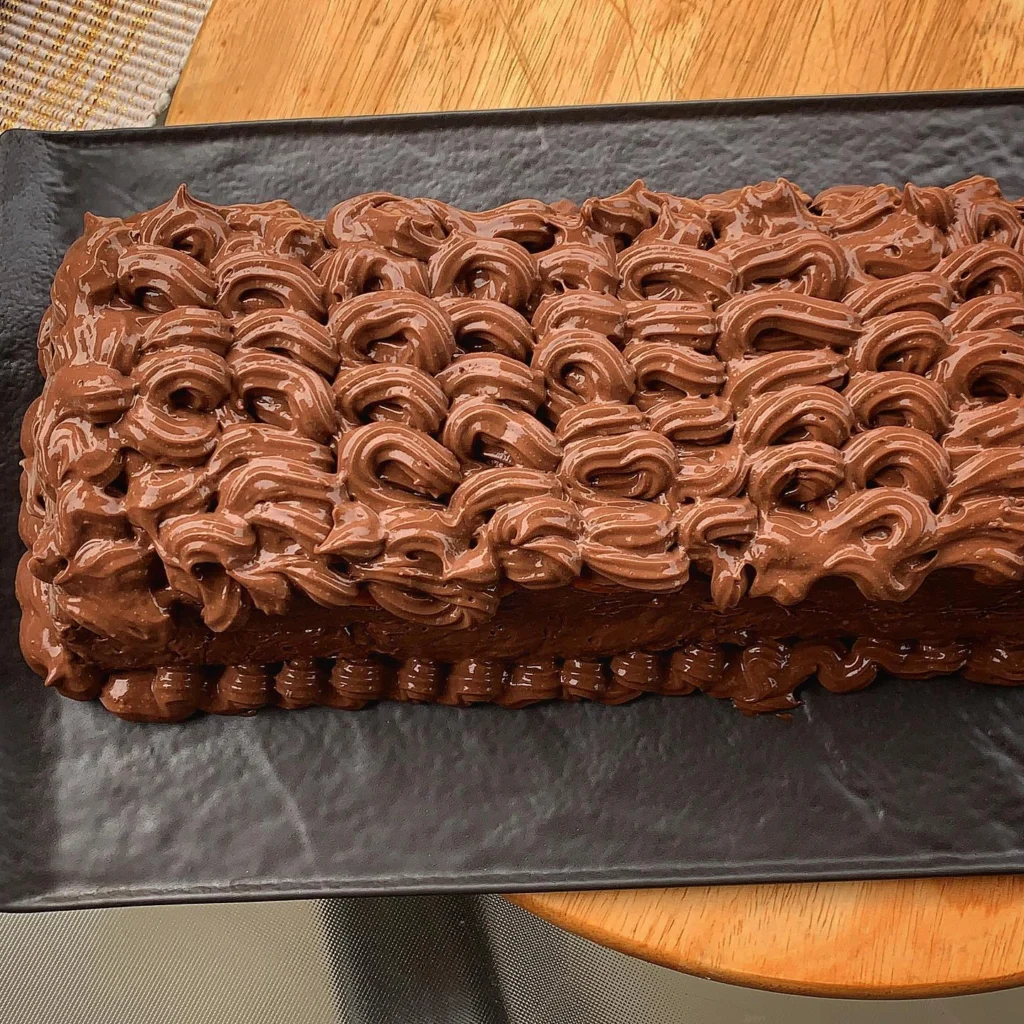 Gotova torta prekrivena valovitim slojem čokoladne kreme, snimljena odozgo na crnom tanjuru s toplim svjetlom i drvenom podlogom