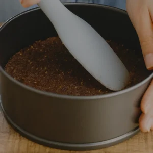Ruka pritiska podlogu od badema i datulja u kalupu za cheesecake pomoću silikonske špatule