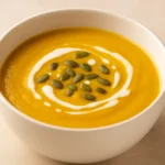 Krem juha od bundeve u bijeloj zdjeli, ukrašena sjemenkama bundeve i kapljicama vrhnja, snimljena u prirodnom svjetlu, savršena za jesenski ručak.
