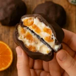 Ruka drži prepolovljeni zdravi čokoladni zalogaj od mandarina i jogurta prekriven tamnom čokoladom, u rustikalnom balkanskom ambijentu, realistična food fotografija bez pečenja.